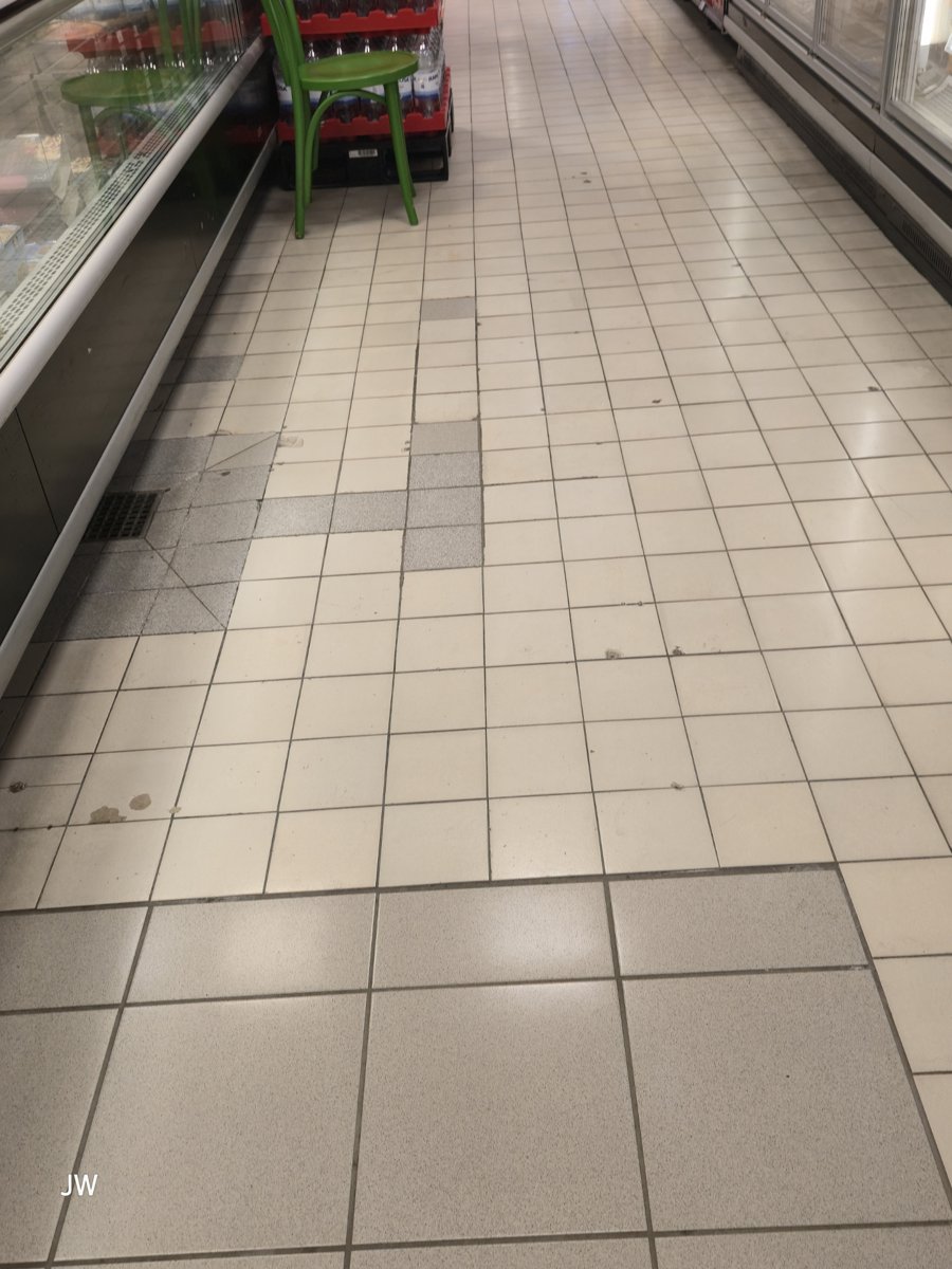 Zdjęcie posadzki z płytek ceramicznych w sklepie spożywczym, z łatami z szarych płytek i kratką odpływową, wysłane do obu asystentów