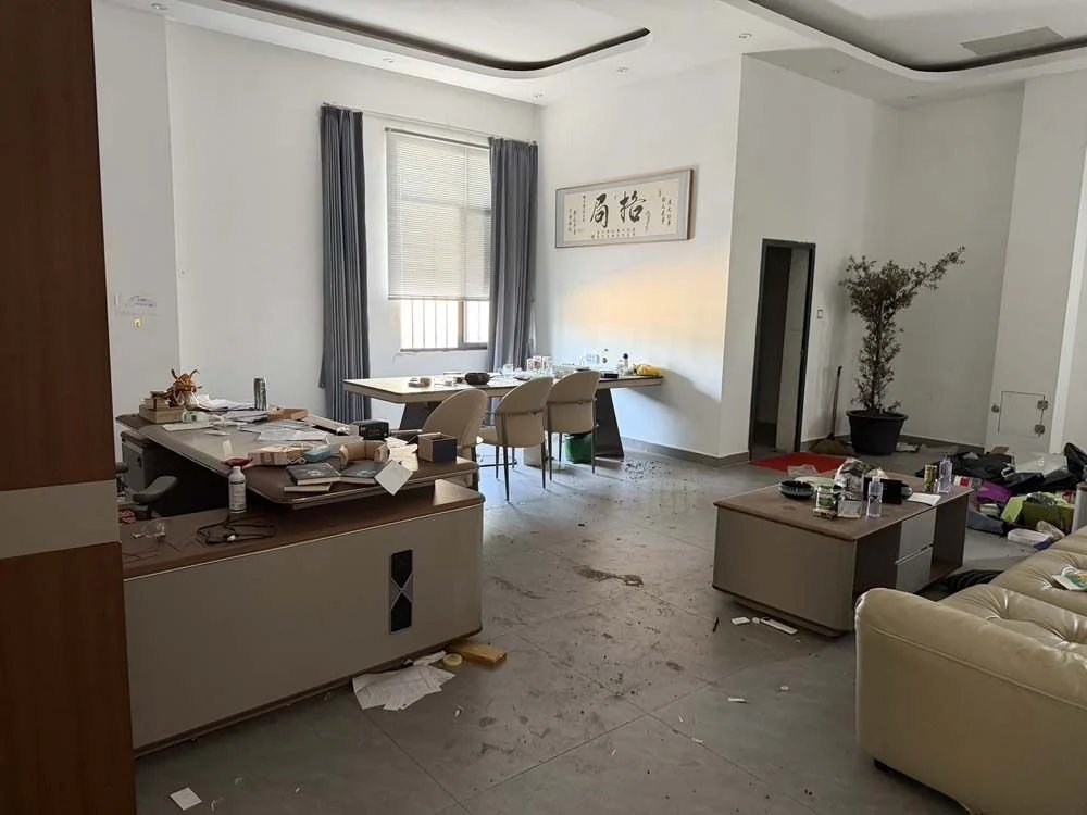 Zdjęcie dużego zabałaganionego pomieszczenia z biurkami i sofą oraz zabrudzoną podłogą z szarych płyt, wysłane do obu asystentów